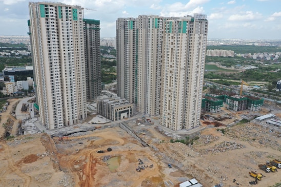 Multiple high-rise residential buildings under construction are surrounded by a mix of cleared land and other construction activities. The area is partly developed with a combination of dirt and concrete surfaces. Nearby, several smaller buildings and construction equipment can be seen, with green netting on some structures indicating an ongoing project.