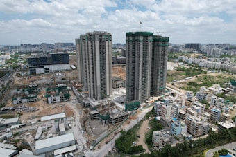 A cityscape featuring high-rise buildings under construction surrounded by various smaller residential buildings. Construction cranes are visible on the site with materials and machinery scattered around. The skyline extends into the distance with additional buildings and a cloudy sky overhead.