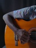 A close-up of a person playing an acoustic guitar with a focus on the right hand strumming the strings. The background is dark, emphasizing the vibrant color of the guitar body and the patterned shirt sleeve.