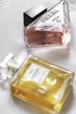 Two perfume bottles are placed on a white surface. One bottle is a triangular glass container with a pink liquid, labeled 'Prada'. The other is a rectangular bottle with a yellow liquid, labeled 'Chanel No. 5'. Both bottles have elegant and classic designs, showcasing luxury brands.