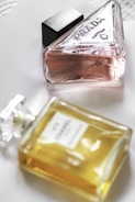 Two perfume bottles are placed on a white surface. One bottle is a triangular glass container with a pink liquid, labeled 'Prada'. The other is a rectangular bottle with a yellow liquid, labeled 'Chanel No. 5'. Both bottles have elegant and classic designs, showcasing luxury brands.