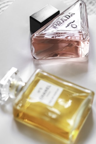 Two perfume bottles are placed on a white surface. One bottle is a triangular glass container with a pink liquid, labeled 'Prada'. The other is a rectangular bottle with a yellow liquid, labeled 'Chanel No. 5'. Both bottles have elegant and classic designs, showcasing luxury brands.