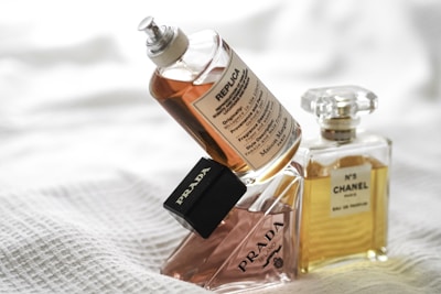 Three perfume bottles are artfully arranged against a soft white textured background. A Replica bottle is on the top, appearing to be in use, next to a stylishly angled Prada bottle, and a classic Chanel No. 5 bottle stands in the background.