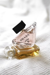 Two perfume bottles are lying on a textured white fabric. One bottle, triangular in shape, is labeled with 'Prada Milano' and stands upright while leaning against the other. The second bottle is rectangular, filled with amber liquid, and labeled 'Chanel No. 5'.