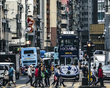 A busy urban street with multiple people crossing the road. There is a prominent tram with the text HKMOA on the front, indicating it is part of the Hong Kong Museum of Art. Several buses and cars are also visible on the street. Surrounding the street are tall, densely packed buildings with numerous signs and advertisements.