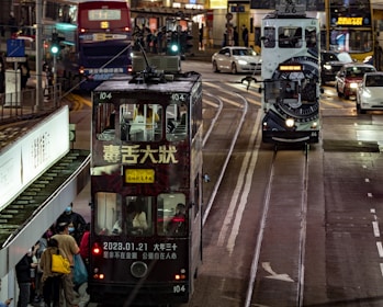 A busy urban street scene with two trams operating on parallel tracks. One tram is painted with various designs, while the other has large text and a date visible on it. People are standing at a tram stop, and vehicles, including a bus and a car, are visible in the background. It is night, and the street is illuminated by artificial lighting.