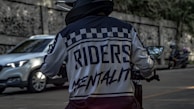 A person in a motorcycle jacket with the words 'Riders Mentality' on the back is riding a motorcycle on a street. The scene includes a blurred white car and two motorcycles in the background parked beside a stone wall.