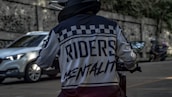 A person in a motorcycle jacket with the words 'Riders Mentality' on the back is riding a motorcycle on a street. The scene includes a blurred white car and two motorcycles in the background parked beside a stone wall.