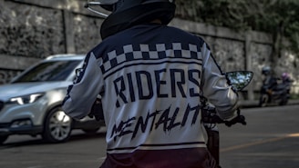 A person in a motorcycle jacket with the words 'Riders Mentality' on the back is riding a motorcycle on a street. The scene includes a blurred white car and two motorcycles in the background parked beside a stone wall.
