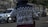 A person in a motorcycle jacket with the words 'Riders Mentality' on the back is riding a motorcycle on a street. The scene includes a blurred white car and two motorcycles in the background parked beside a stone wall.