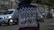 A person in a motorcycle jacket with the words 'Riders Mentality' on the back is riding a motorcycle on a street. The scene includes a blurred white car and two motorcycles in the background parked beside a stone wall.