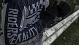 A person wearing a black and white long-sleeve shirt with the words 'RIDERS' and 'MENTALITY' printed on it stands next to a metal barrier. The individual is adjusting or wearing black gloves. The setting appears to be outdoors, with some greenery visible in the background.