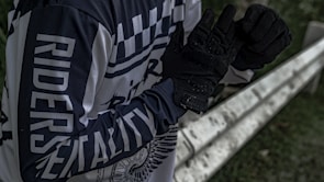 A person wearing a black and white long-sleeve shirt with the words 'RIDERS' and 'MENTALITY' printed on it stands next to a metal barrier. The individual is adjusting or wearing black gloves. The setting appears to be outdoors, with some greenery visible in the background.