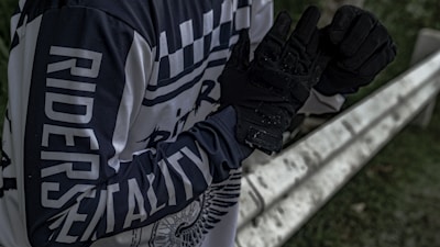 A person wearing a black and white long-sleeve shirt with the words 'RIDERS' and 'MENTALITY' printed on it stands next to a metal barrier. The individual is adjusting or wearing black gloves. The setting appears to be outdoors, with some greenery visible in the background.