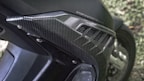 A close-up view of a motorcycle part, featuring a carbon fiber pattern on a body panel. The detailing includes a textured design with several linear cuts serving as venting. The text 'STOP & START' is partially visible, indicating a feature or branding. Rivets secure the panel in place, showcasing mechanical elements typical of motorcycle construction. The background includes a grassy area that is out of focus.