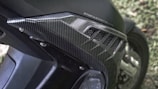 A close-up view of a motorcycle part, featuring a carbon fiber pattern on a body panel. The detailing includes a textured design with several linear cuts serving as venting. The text 'STOP & START' is partially visible, indicating a feature or branding. Rivets secure the panel in place, showcasing mechanical elements typical of motorcycle construction. The background includes a grassy area that is out of focus.
