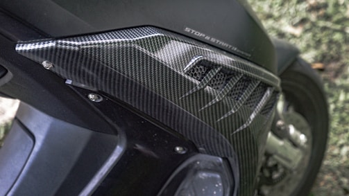 A close-up view of a motorcycle part, featuring a carbon fiber pattern on a body panel. The detailing includes a textured design with several linear cuts serving as venting. The text 'STOP & START' is partially visible, indicating a feature or branding. Rivets secure the panel in place, showcasing mechanical elements typical of motorcycle construction. The background includes a grassy area that is out of focus.