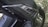 A close-up view of a motorcycle part, featuring a carbon fiber pattern on a body panel. The detailing includes a textured design with several linear cuts serving as venting. The text 'STOP & START' is partially visible, indicating a feature or branding. Rivets secure the panel in place, showcasing mechanical elements typical of motorcycle construction. The background includes a grassy area that is out of focus.