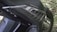 A close-up view of a motorcycle part, featuring a carbon fiber pattern on a body panel. The detailing includes a textured design with several linear cuts serving as venting. The text 'STOP & START' is partially visible, indicating a feature or branding. Rivets secure the panel in place, showcasing mechanical elements typical of motorcycle construction. The background includes a grassy area that is out of focus.