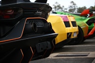 The image features the rear view of three high-performance sports cars parked in a row. The foreground shows a black car with orange detailing around the exhaust. Beside it is a vibrant yellow car, followed by a green car with red accents and intricate patterns. The background is slightly blurred with trees visible in the distance.