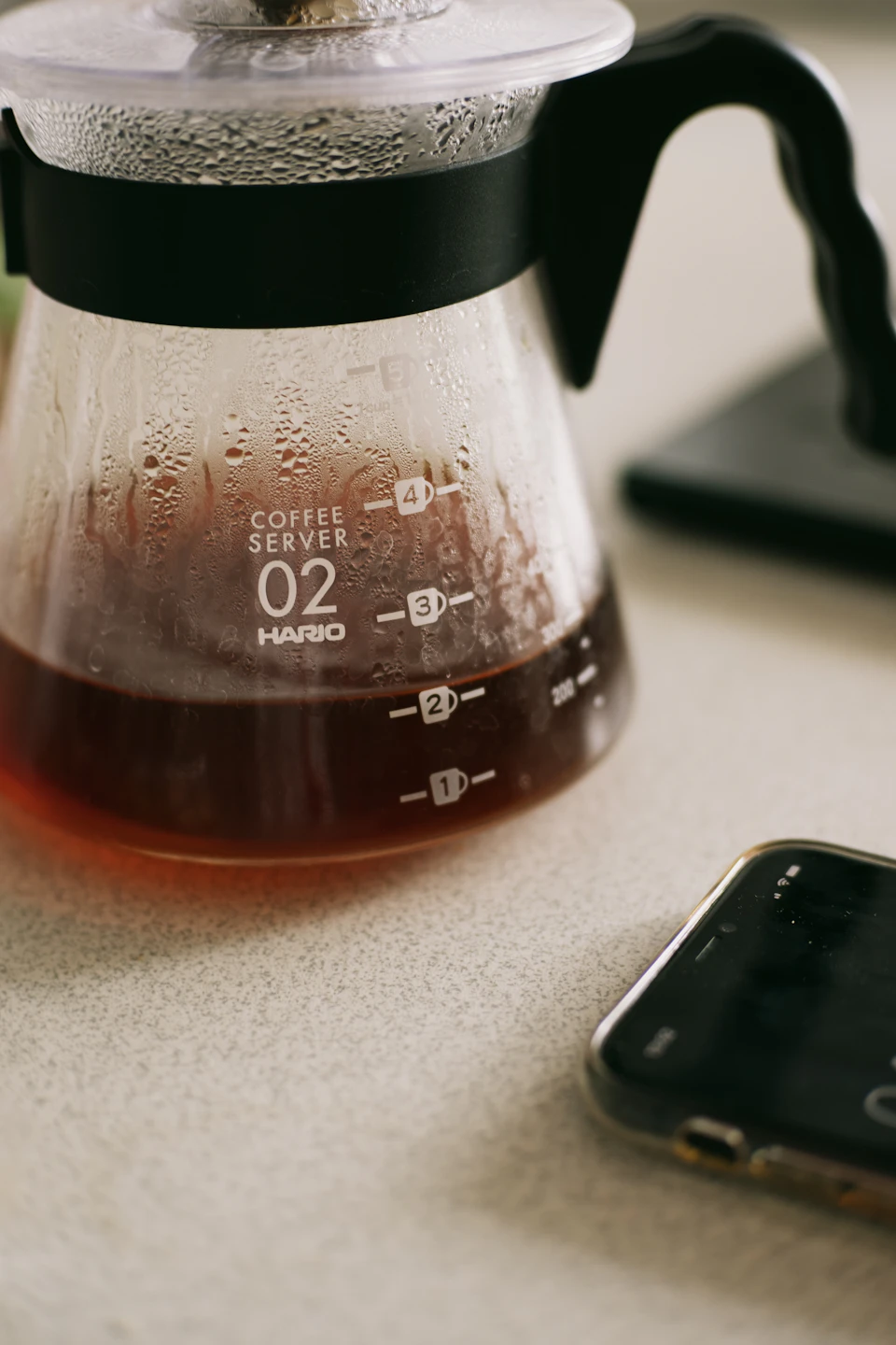 The image shows a close-up view of a coffee maker on a white countertop, with a black handle and spout. The coffee maker is filled to the brim with dark liquid, indicating it has been freshly brewed. A cell phone lies next to the coffee maker, suggesting that someone may have recently used or is about to use it for communication or other tasks.