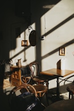 A small cafe setting with wooden chairs and tables. Sunlight streams through a window, casting long shadows across the room. On the wall, small framed pictures and a circular object are visible, adding a cozy and artistic touch. The scene appears tranquil with muted tones.