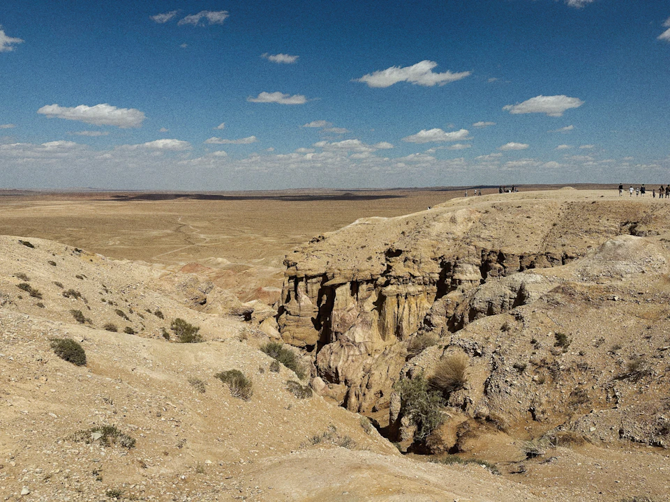 The image depicts a desert landscape with a large, jagged cliff face on the right side and a flat, sandy plain extending towards the left side. The sky above is clear blue with scattered clouds. In the distance, several people can be seen walking along the edge of the cliff, adding a sense of scale to the scene.

The image does not contain any discernible text or countable objects, but it captures the vastness and rugged beauty of the desert landscape.