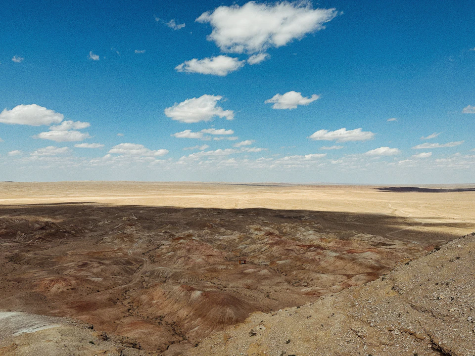 The image depicts a vast, barren desert landscape under a clear blue sky with fluffy white clouds. The ground is covered in dry, brown sand that stretches out to meet the horizon at the edge of the frame. In the distance, there are small hills and mountains visible against the backdrop of the expansive desert.