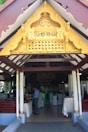 An ornate golden gable decorates the entrance of a pavilion with white columns and wooden beams. Inside, people are gathered, some serving themselves from a buffet, with tables draped in white cloths. The setting suggests an event or gathering.