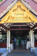 An ornate golden gable decorates the entrance of a pavilion with white columns and wooden beams. Inside, people are gathered, some serving themselves from a buffet, with tables draped in white cloths. The setting suggests an event or gathering.