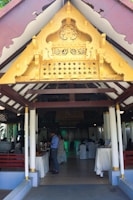 An ornate golden gable decorates the entrance of a pavilion with white columns and wooden beams. Inside, people are gathered, some serving themselves from a buffet, with tables draped in white cloths. The setting suggests an event or gathering.