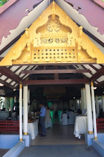 An ornate golden gable decorates the entrance of a pavilion with white columns and wooden beams. Inside, people are gathered, some serving themselves from a buffet, with tables draped in white cloths. The setting suggests an event or gathering.