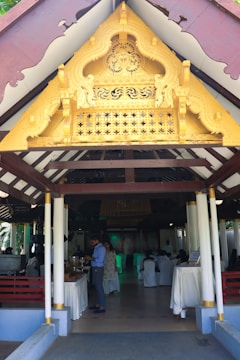 An ornate golden gable decorates the entrance of a pavilion with white columns and wooden beams. Inside, people are gathered, some serving themselves from a buffet, with tables draped in white cloths. The setting suggests an event or gathering.