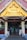 An ornate golden gable decorates the entrance of a pavilion with white columns and wooden beams. Inside, people are gathered, some serving themselves from a buffet, with tables draped in white cloths. The setting suggests an event or gathering.