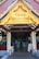 An ornate golden gable decorates the entrance of a pavilion with white columns and wooden beams. Inside, people are gathered, some serving themselves from a buffet, with tables draped in white cloths. The setting suggests an event or gathering.