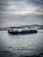 A ferry is navigating calm waters with a city skyline in the background. The sky is overcast with scattered clouds, and the water reflects shades of gray. The ferry is painted in white with blue and red accents, and a Turkish flag is visible.