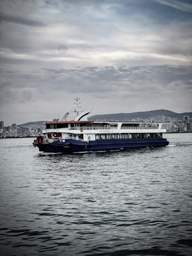 A ferry is navigating calm waters with a city skyline in the background. The sky is overcast with scattered clouds, and the water reflects shades of gray. The ferry is painted in white with blue and red accents, and a Turkish flag is visible.