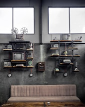 A vintage-themed room with industrial decor, featuring shelves holding old electronic devices including televisions, film reels, radios, typewriters, and a rotary telephone. The walls are concrete grey, and two large windows add natural light to the setting. A beige cushioned bench and a rustic wooden table are positioned below the shelves.