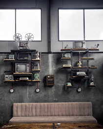 A vintage-themed room with industrial decor, featuring shelves holding old electronic devices including televisions, film reels, radios, typewriters, and a rotary telephone. The walls are concrete grey, and two large windows add natural light to the setting. A beige cushioned bench and a rustic wooden table are positioned below the shelves.