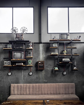 A vintage-themed room with industrial decor, featuring shelves holding old electronic devices including televisions, film reels, radios, typewriters, and a rotary telephone. The walls are concrete grey, and two large windows add natural light to the setting. A beige cushioned bench and a rustic wooden table are positioned below the shelves.