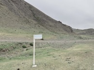 A barren landscape with a rugged hillside and sparse vegetation. A signpost in the foreground has a message in two languages reminding people to 'Keep save natural.' The colors are muted, with shades of brown and green dominating the scene.