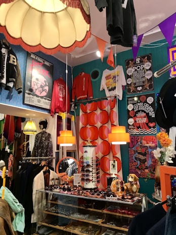 A vibrant and eclectic boutique showcasing a mix of vintage and retro items. The space is decorated with colorful posters and clothing on display, including jackets and T-shirts hanging from the ceiling and walls. A glass display case contains an assortment of sunglasses, with orange pendant lamps and bold, patterned wallpaper adding to the lively atmosphere. A quirky collection of items, such as a dog figurine and decorative flowers, enhances the playful and nostalgic feel.