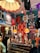 A vibrant and eclectic boutique showcasing a mix of vintage and retro items. The space is decorated with colorful posters and clothing on display, including jackets and T-shirts hanging from the ceiling and walls. A glass display case contains an assortment of sunglasses, with orange pendant lamps and bold, patterned wallpaper adding to the lively atmosphere. A quirky collection of items, such as a dog figurine and decorative flowers, enhances the playful and nostalgic feel.