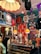 A vibrant and eclectic boutique showcasing a mix of vintage and retro items. The space is decorated with colorful posters and clothing on display, including jackets and T-shirts hanging from the ceiling and walls. A glass display case contains an assortment of sunglasses, with orange pendant lamps and bold, patterned wallpaper adding to the lively atmosphere. A quirky collection of items, such as a dog figurine and decorative flowers, enhances the playful and nostalgic feel.