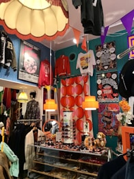 A vibrant and eclectic boutique showcasing a mix of vintage and retro items. The space is decorated with colorful posters and clothing on display, including jackets and T-shirts hanging from the ceiling and walls. A glass display case contains an assortment of sunglasses, with orange pendant lamps and bold, patterned wallpaper adding to the lively atmosphere. A quirky collection of items, such as a dog figurine and decorative flowers, enhances the playful and nostalgic feel.