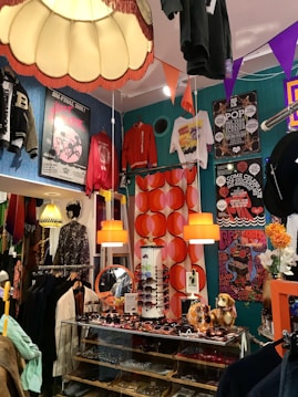 A vibrant and eclectic boutique showcasing a mix of vintage and retro items. The space is decorated with colorful posters and clothing on display, including jackets and T-shirts hanging from the ceiling and walls. A glass display case contains an assortment of sunglasses, with orange pendant lamps and bold, patterned wallpaper adding to the lively atmosphere. A quirky collection of items, such as a dog figurine and decorative flowers, enhances the playful and nostalgic feel.