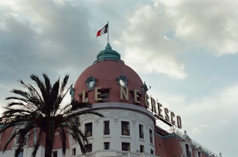 A prominent historical building with a dome and the French flag on top. It features a large sign with the letters 'LE NEGRESCO'. There are classical architectural elements and a palm tree is visible to the left.