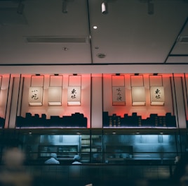 A dimly lit interior with traditional lanterns adorned with Chinese calligraphy hanging on a wall. Below, a kitchen area is visible through glass, where chefs, wearing white uniforms and hats, are focused on their culinary tasks.