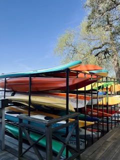 A stack of colorful kayaks is organized on a wooden rack. The kayaks are in various shades such as orange, yellow, and blue. A large tree with green leaves is in the background under a clear blue sky.