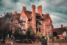 A large, red-brick building with prominent chimneys stands under a partly cloudy sky. The architecture is traditional, with gabled roofs and detailed brickwork. Surrounding the building are mature trees and neatly trimmed hedges. In the foreground, a person wearing a dark top and blue jeans is standing near a low brick wall, next to a street sign labeled 'PRINCESS ROAD EAST.'